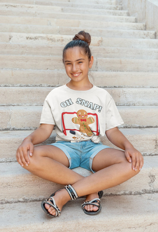 Girl sitting on stairs wearing a white t-shirt. It says oh snap with the graphic of a gingerbread man with a broken leg in front of a hockey net. There is a puck in front.