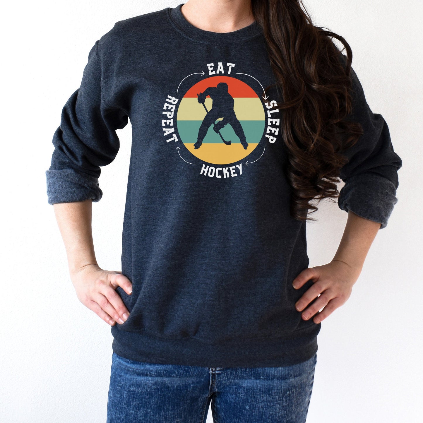 Woman wearing a dark heather Gildan 18000 unisex crewneck sweatshirt. It says eat, sleep, hockey, repeat in the circle with arrows between each word. There is a retro style striped sunset in the middle with the cutout of a hockey player.