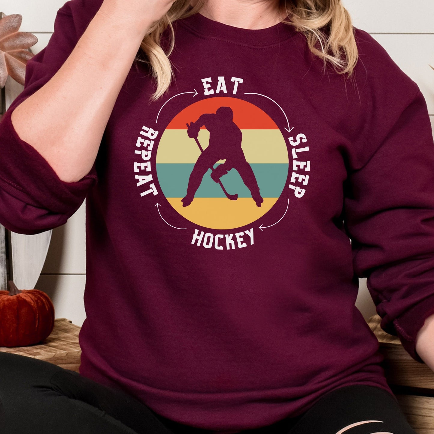 Woman wearing a maroon Gildan 18000 unisex crewneck sweatshirt. It says eat, sleep, hockey, repeat in the circle with arrows between each word. There is a retro style striped sunset in the middle with the cutout of a hockey player.