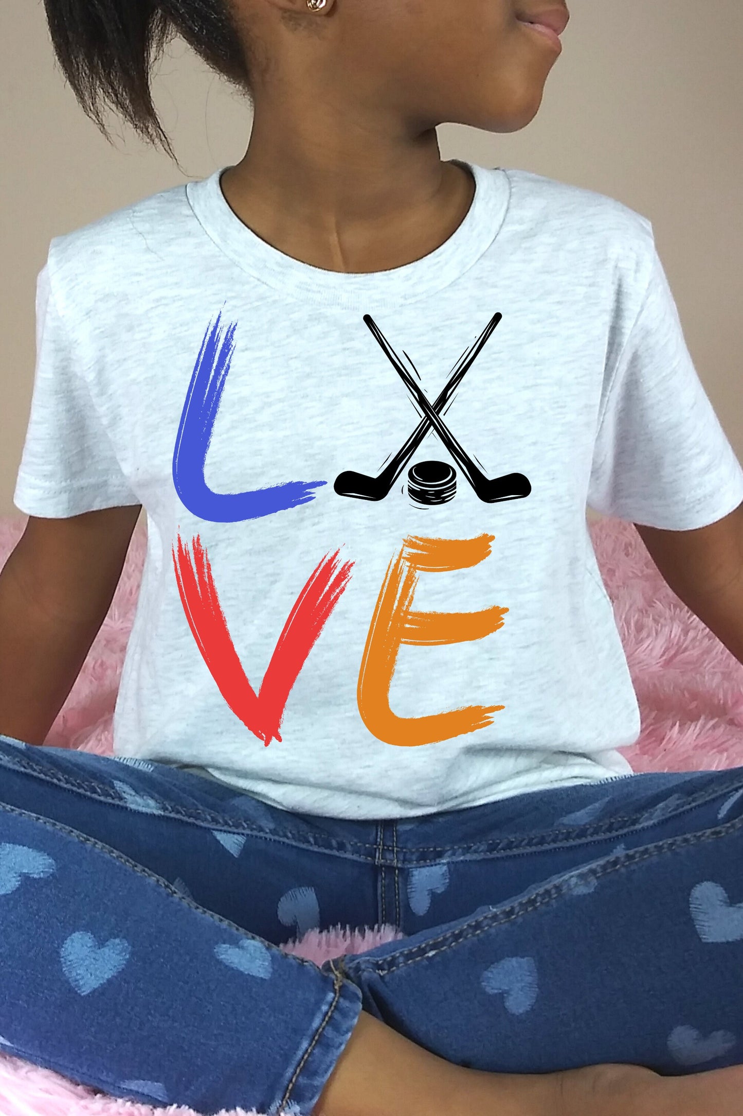 Girl wearing an ash Bella and Canvas 3001Y crewneck youth t-shirt. It says love in a brushed paint font. The o is replaced with two crossed hockey sticks and a hockey puck.