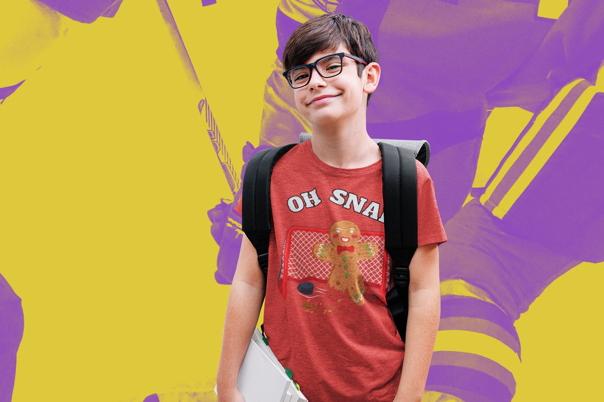 Boy wearing a heather red t-shirt. It says oh snap with the graphic of a gingerbread man with a broken leg in front of a hockey net. There is a puck in front.