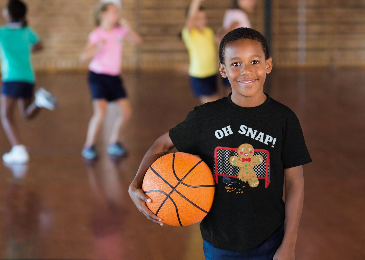 Boy wearing a black t-shirt holding a basketball. The shirt says oh snap. There is a graphic of a gingerbread man with a broken leg in front of a hockey net.