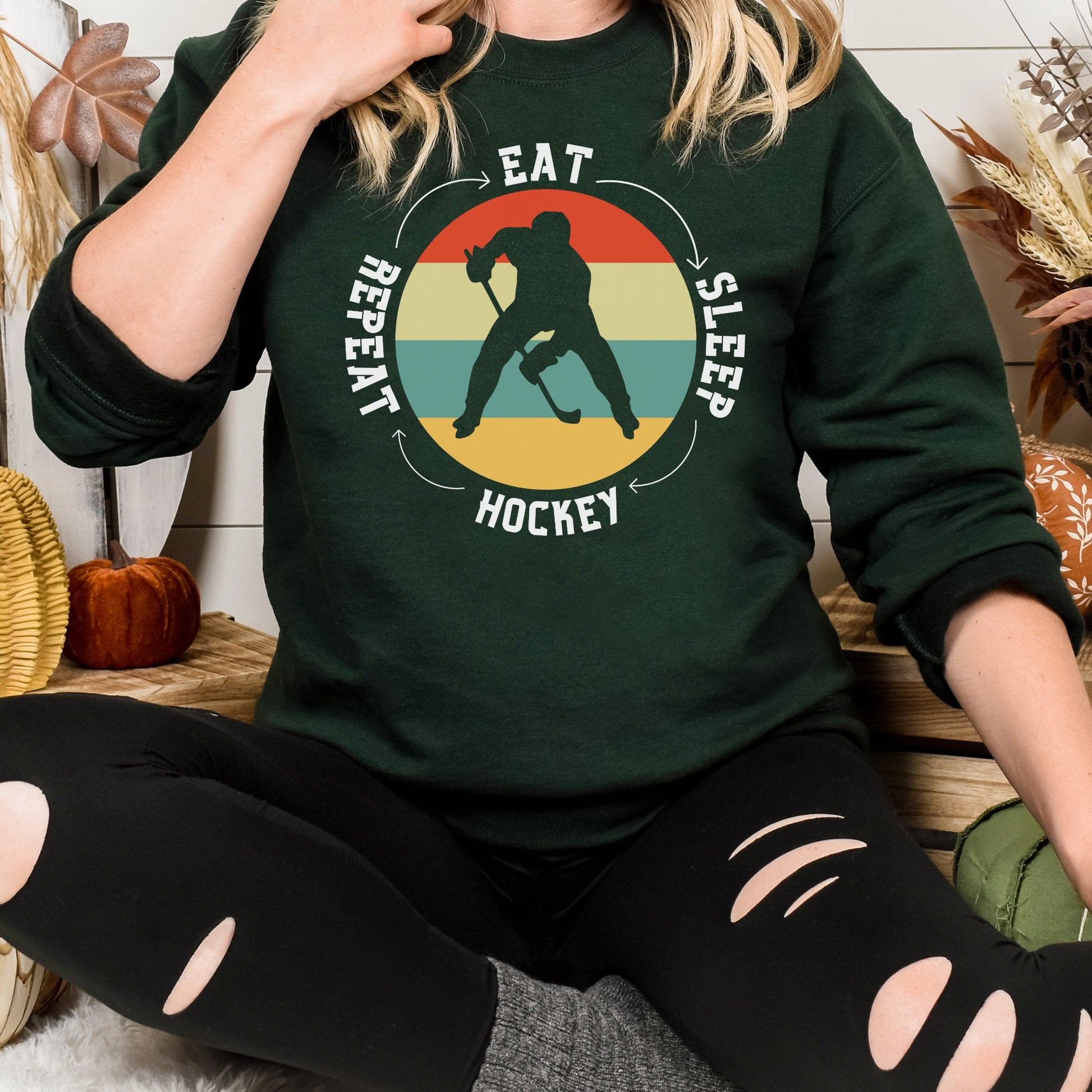 Woman wearing a forest Gildan 18000 unisex crewneck sweatshirt. It says eat, sleep, hockey, repeat in the circle with arrows between each word. There is a retro style striped sunset in the middle with the cutout of a hockey player.