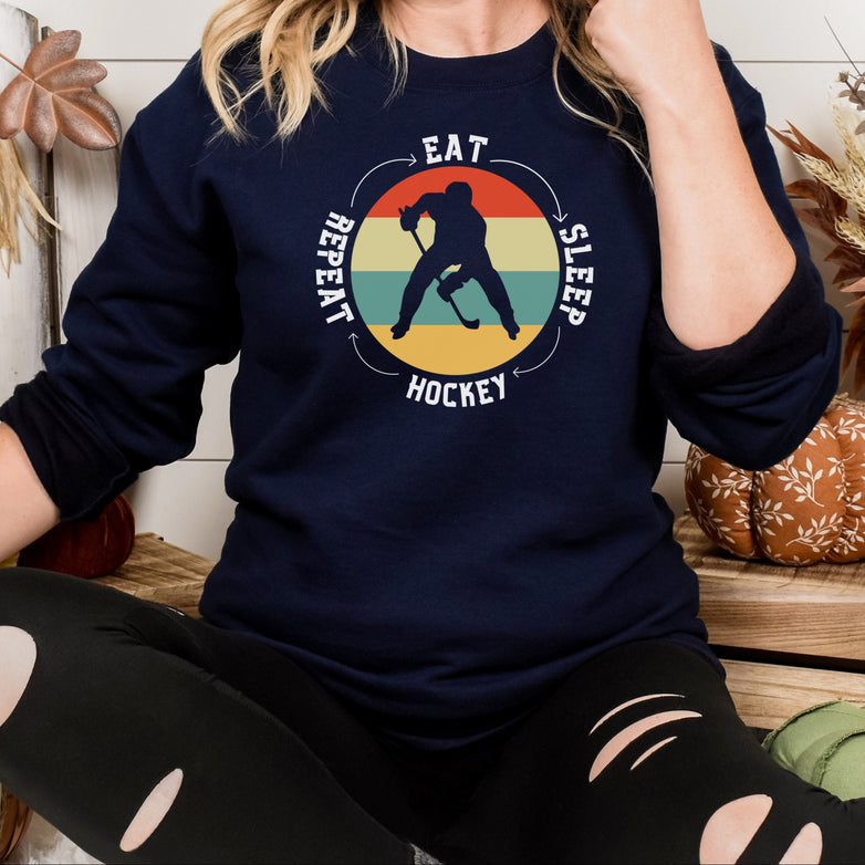 Woman wearing a navy Gildan 18000 unisex crewneck sweatshirt. It says eat, sleep, hockey, repeat in the circle with arrows between each word. There is a retro style striped sunset in the middle with the cutout of a hockey player.