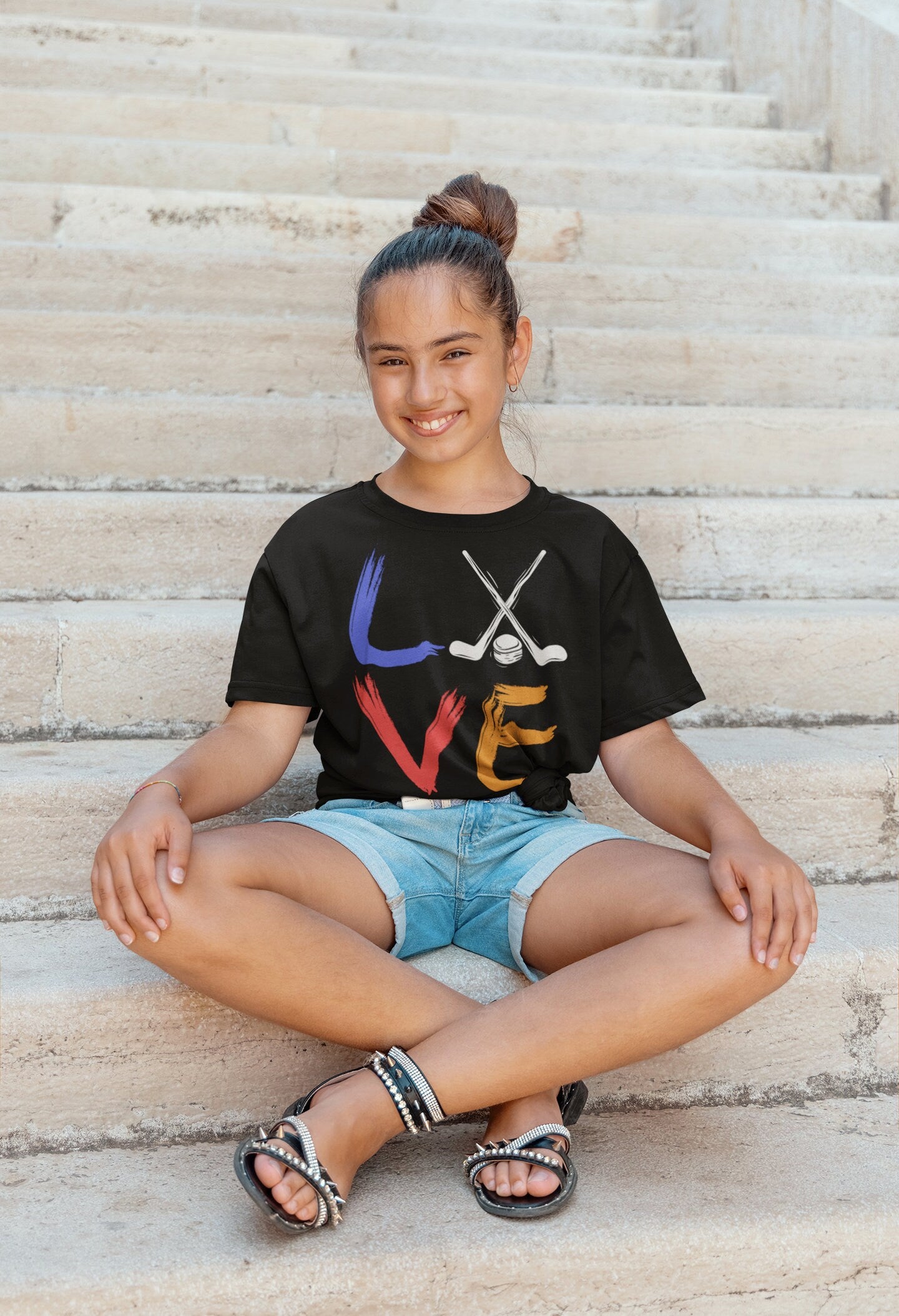 Girl wearing a heather black Bella and Canvas 3001Y crewneck youth t-shirt. It says love in a brushed paint font. The o is replaced with two crossed hockey sticks and a hockey puck.