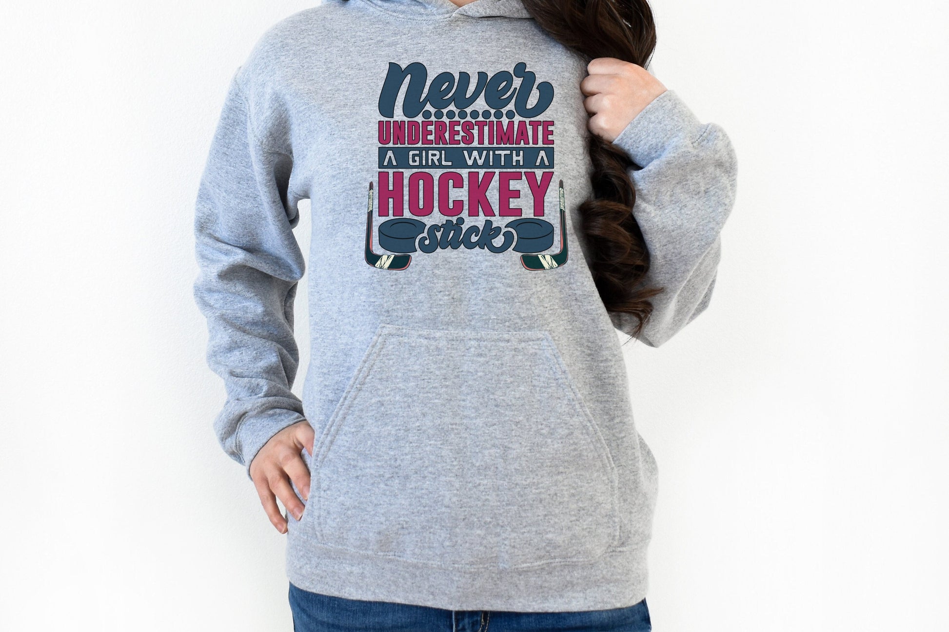 Sport grey Gildan 18500 unisex crewneck sweatshirt. It says never underestimate a girl with a hockey stick in dark blue and hot pink. There are two pucks beside the word stick. There are hockey sticks on either side of the text.