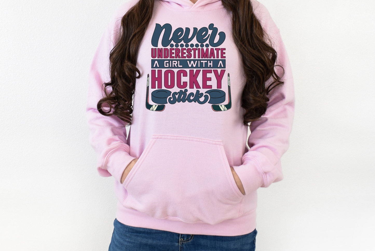 Light pink Gildan 18500 unisex crewneck sweatshirt. It says never underestimate a girl with a hockey stick in dark blue and hot pink. There are two pucks beside the word stick. There are hockey sticks on either side of the text.