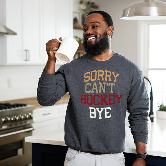 Smiling man holding a coffee mug wearing a dark heather Gildan 18000 unisex crewneck sweatshirt. It says sorry cant hockey bye in different colored font. The words are bold and lined up vertically.