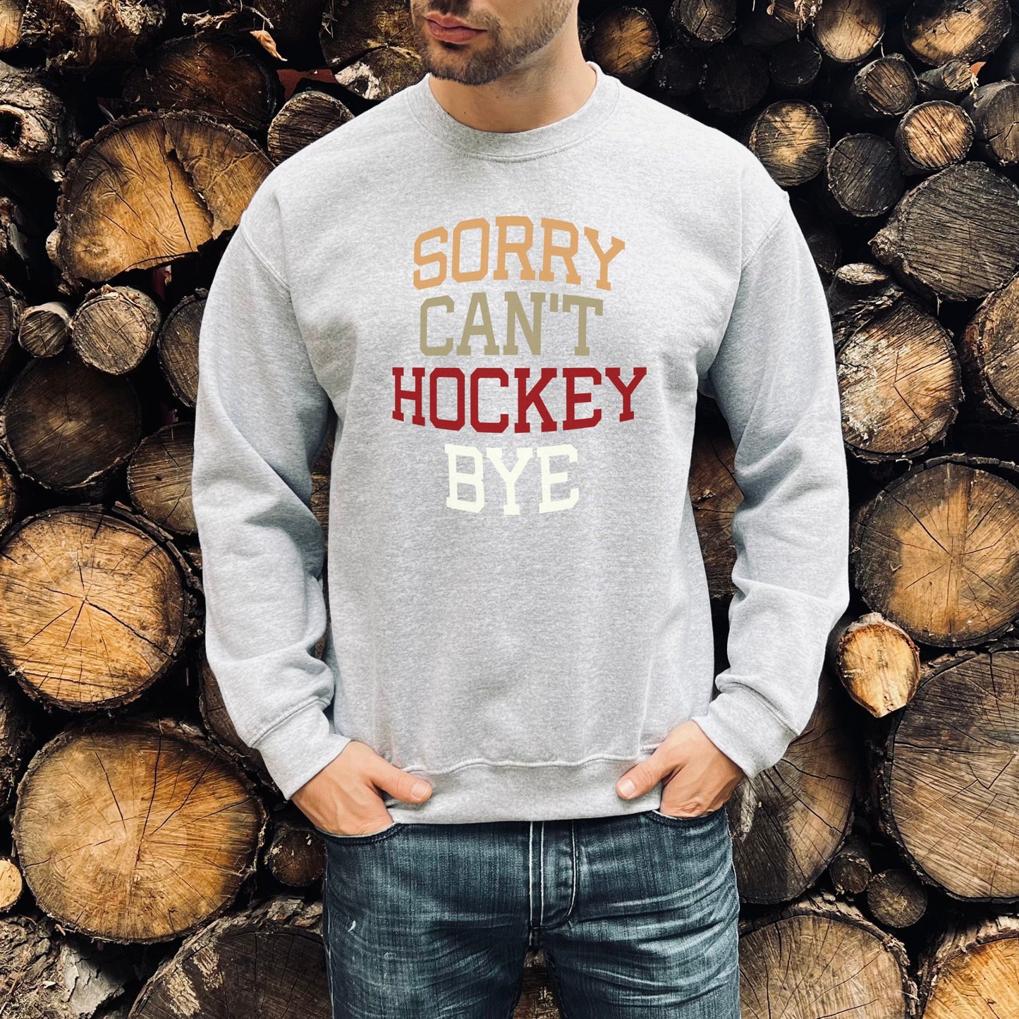 Man standing in front of a pile of logs wearing a sport grey Gildan 18000 unisex crewneck sweatshirt. It says sorry cant hockey bye in different colored font. The words are bold and lined up vertically.