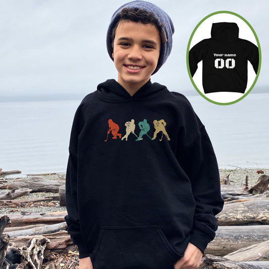 Boy wearing a black Gildan 18500B youth hoodie. It has four figures of hockey players in a vintage distressed style. The colors are red, cream, blue and yellow. Personalized name and number available on the back.