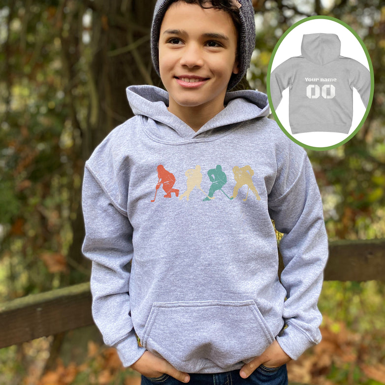 Boy wearing a sport grey Gildan 18500B youth hoodie. It has four figures of hockey players in a vintage distressed style. The colors are red, cream, blue and yellow. Personalized name and number available on the back.