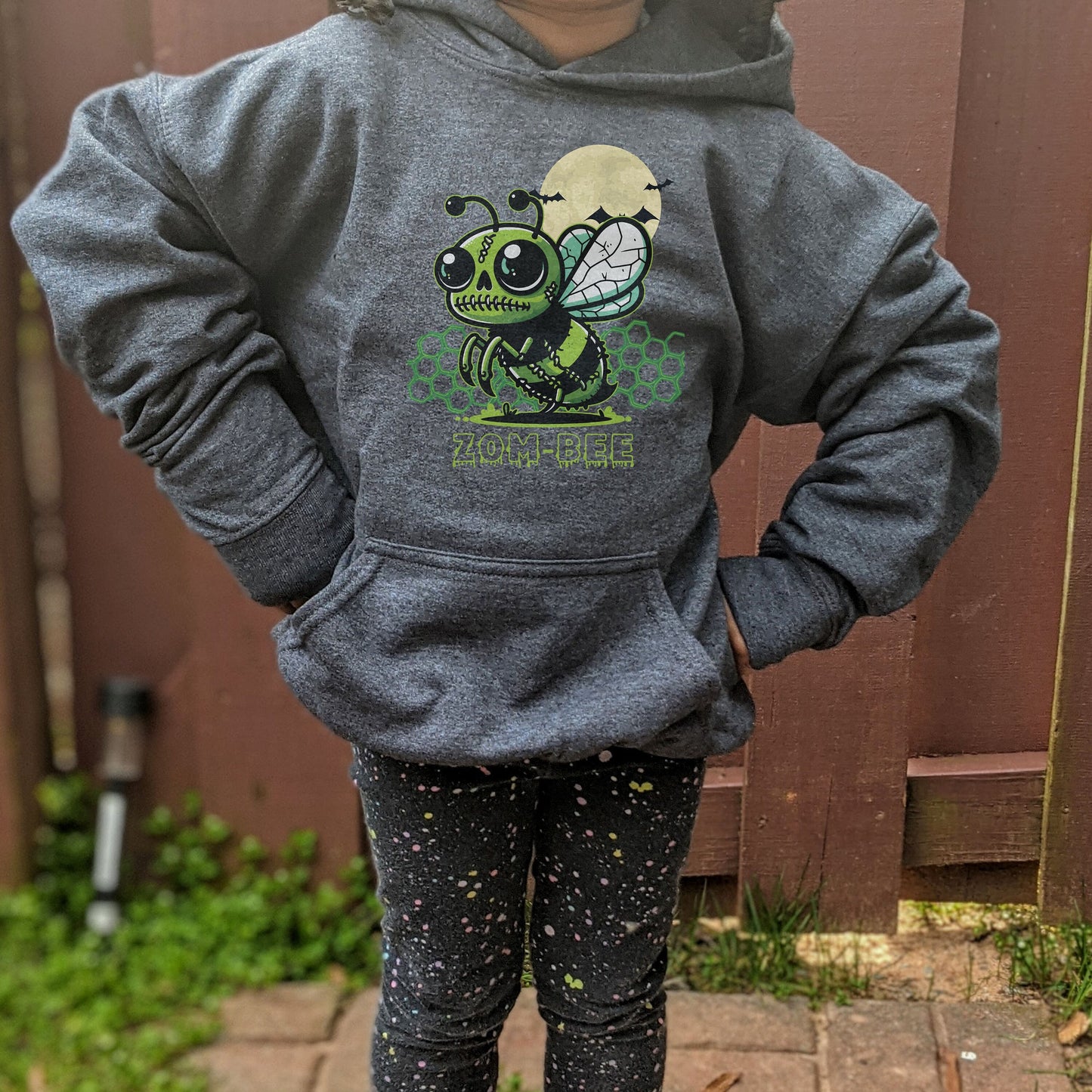 Girl wearing a dark heather Gildan 18500B youth hoodie. It has a drawing of a green and black zombie bee in the middle with a moon with bats and green honeycomb behind it. It says zom-bee at the bottom in a green drippy font.