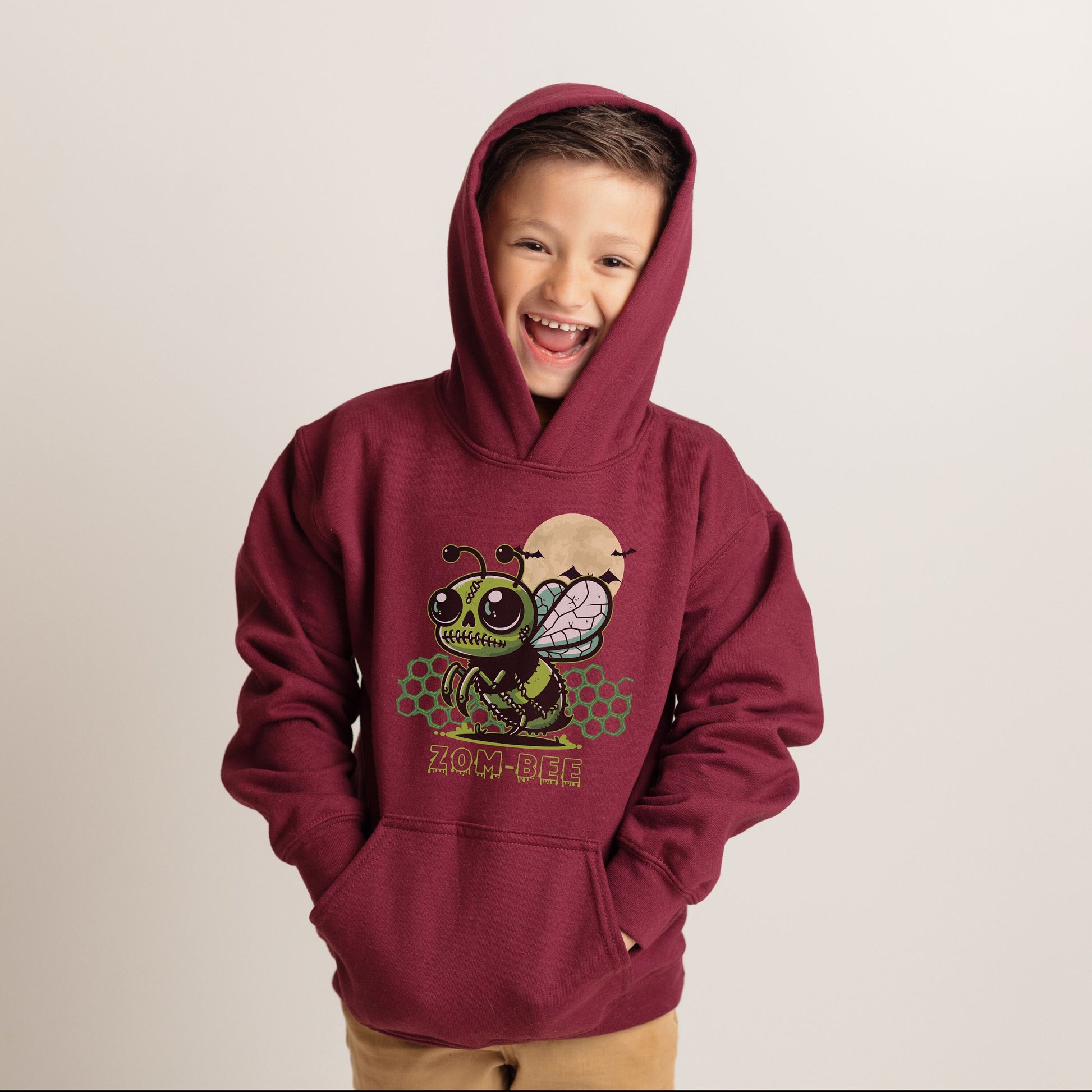 Boy wearing a maroon Gildan 18500B youth hoodie. It has a drawing of a green and black zombie bee in the middle with a moon with bats and green honeycomb behind it. It says zom-bee at the bottom in a green drippy font.