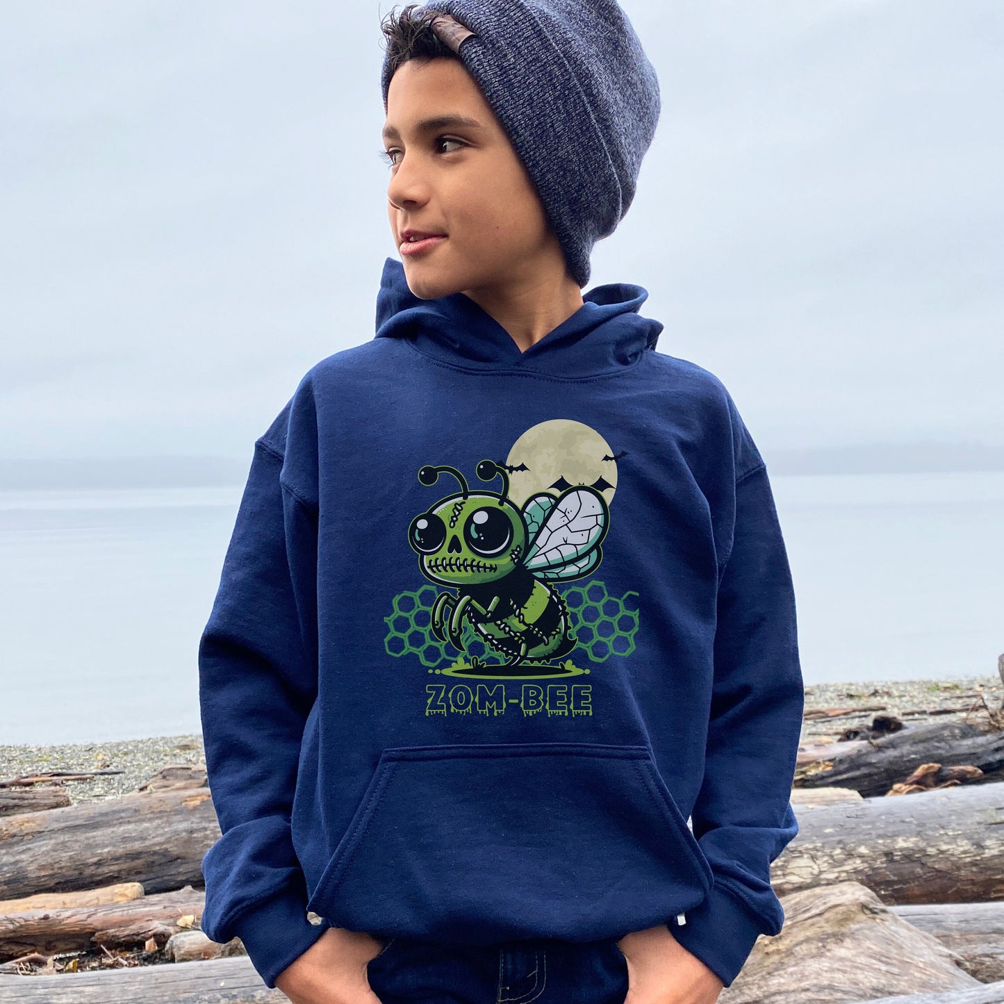 Boy wearing a navy Gildan 18500B youth hoodie. It has a drawing of a green and black zombie bee in the middle with a moon with bats and green honeycomb behind it. It says zom-bee at the bottom in a green drippy font.